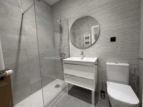Baño moderno en habitación individual MTM Rooms, Santander con ducha abierta, lavabo blanco, espejo redondo e inodoro