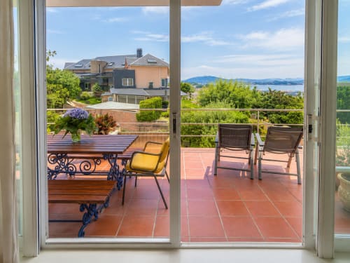 Terraza con vista al mar en Baiona, España. Mesa de madera con flores, silla amarilla y tumbonas con vistas al paisaje costero.