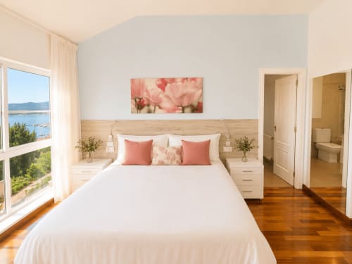 Dormitorio luminoso en cabaña de Baiona con vistas al mar, cabecero de madera, almohadas rosas y ventana con vista al agua