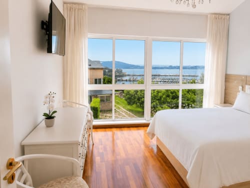 Dormitorio luminoso con cama doble, suelo de madera, amplios ventanales con vistas al mar hacia la marina de Baiona y montañas