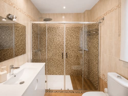 Baño moderno con ducha de azulejos mosaico, lavabo blanco, inodoro y suelo de madera en la casita Alborada de Baiona