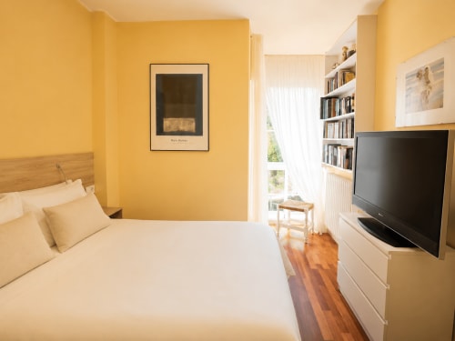 Dormitorio amarillo brillante en casa de Baiona con cama, TV de pantalla plana, estantes, suelos de madera y ventana con vistas al mar