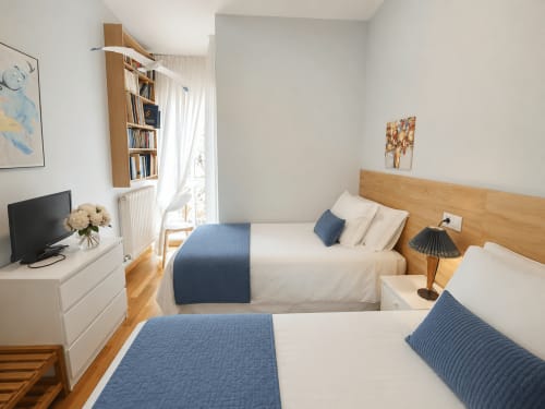 Habitación moderna con dos camas, acentos azules, cabecero de madera, televisor, estantería y vistas al mar en la casita de Baiona