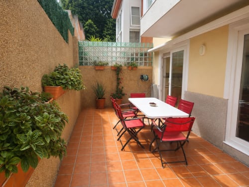 Terraza soleada con sillas rojas, mesa de comedor blanca, plantas en macetas y edificio amarillo en la casa rural Holibai de Baiona