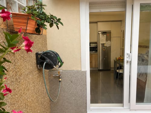 Entrada de casa rural Baiona con plantas en flor, ducha exterior y vista de la cocina a través de puertas de cristal