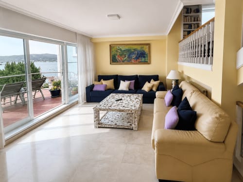 Luminoso salón familiar en amarillo brillante en la casa de Baiona con vistas al mar, sofás en negro y crema, decoración costera y acceso a terraza