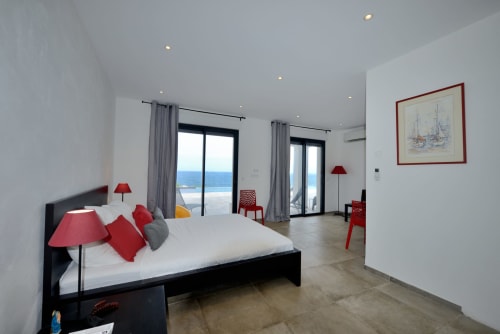Chambre moderne de la Villa EOLE à Centuri en Corse avec vue sur mer, lit noir, touches rouges, rideaux gris et portes-fenêtres vers balcon