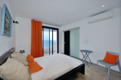 Chambre moderne de la Villa EOLE, Centuri Corse, avec vue mer, lit blanc, touches orangées et portes-fenêtres sur balcon