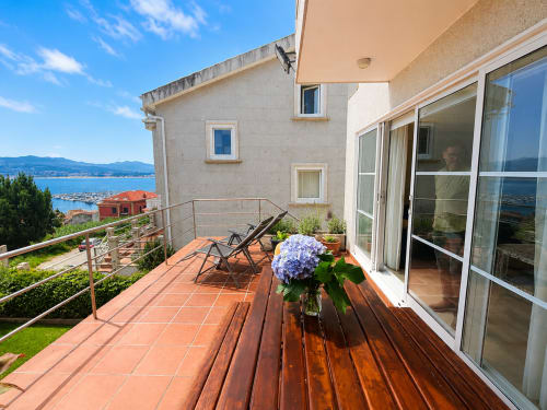Terraza de cabaña en Baiona con vistas al mar, flores azules, tumbonas y bahía al fondo.