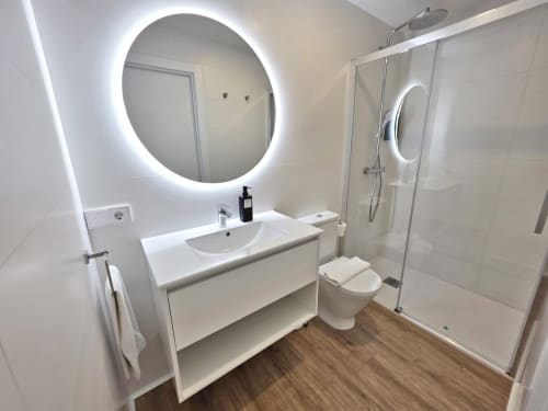 Baño moderno en BR Silgar Sanxenxo con espejo redondo iluminado LED, tocador blanco, inodoro y cabina de ducha de cristal