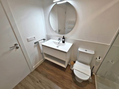 Baño moderno en BR Silgar Sanxenxo con vanidad blanca, espejo ovalado, inodoro y pisos de madera