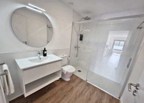 Baño moderno en BR Silgar Sanxenxo con tocador blanco, espejo iluminado redondo, ducha de cristal y piso efecto madera.