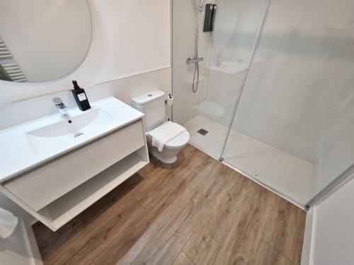 Baño moderno del apartamento BR Silgar Sanxenxo con lavamanos blanco, inodoro, ducha de cristal, suelos de madera y espejo