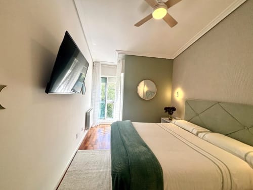 Dormitorio moderno en la casita Serenidade, Baiona con vista al mar, pared verde, TV montada, ventilador de techo y cama confortable