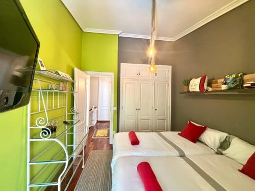 Dormitorio luminoso con paredes verde lima y gris, dos camas con almohadas rojas, TV, estanterías y armarios blancos en la casita Baiona