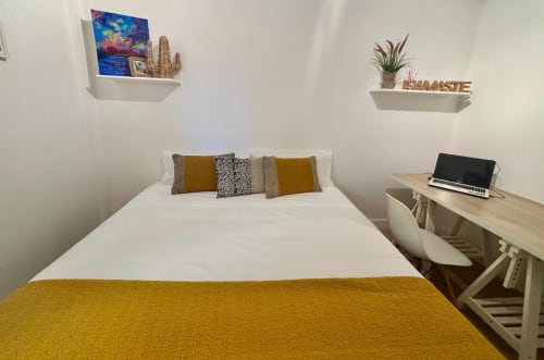 Dormitorio moderno en cabaña de Baiona con paredes blancas, manta amarillo mostaza, almohadas estampadas y escritorio de madera con laptop