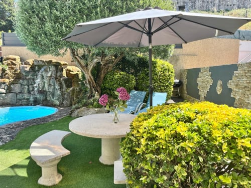 Lujoso patio ajardinado con piscina, sombrilla, asientos de piedra, plantas floridas y construcciones de piedra en la casa rural Holibai de Baiona
