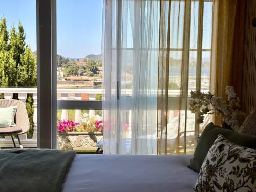 Dormitorio luminoso con vistas al mar en casa de Baiona, cortinas translúcidas, cama con almohadas, balcón, plantas en flor y montañas