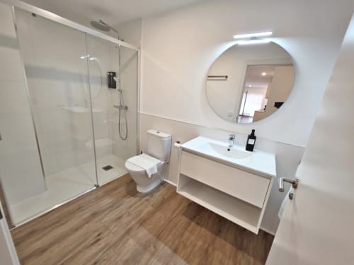 Baño moderno en apartamento BR Silgar, Sanxenxo con ducha de cristal, tocador blanco, espejo redondo y pisos de madera