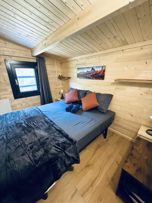 Chambre de lodge moderne en bois avec lit queen, literie bleue, oreillers orange, paysage montagneux et fenêtre à cadre noir