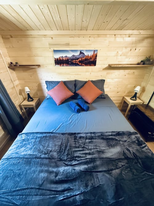 Chambre confortable d'un chalet en bois avec literie bleue, coussins roses et orangés, paysage montagneux et tables de nuit en bois