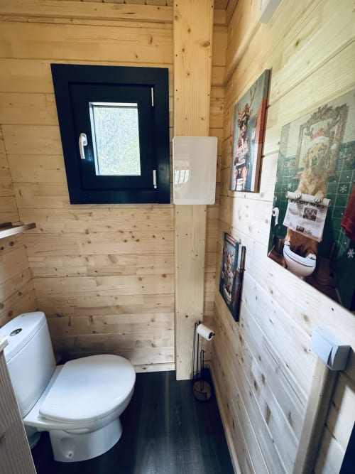 Salle de bain de lodge moderne en bois avec toilettes blanches, sol foncé et fenêtre bleu marine aux Cabanes et lodges du Belvédère