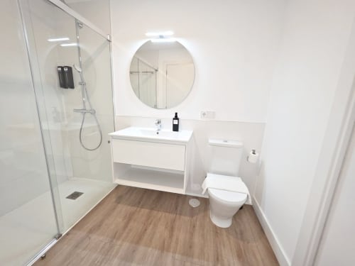 Baño moderno del hotel BR SILGAR en Sanxenxo con ducha abierta, lavamanos blanco, espejo redondo y suelos con aspecto de madera