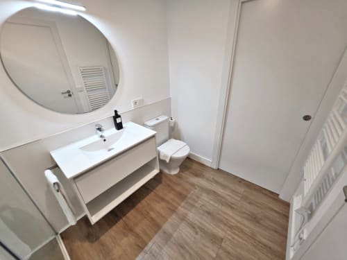 Baño moderno con tocador blanco, espejo redondo, inodoro y suelo con acabado madera en el hotel BR SILGAR de Sanxenxo
