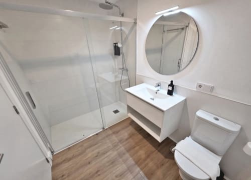 Baño moderno en BR SILGAR Sanxenxo con ducha tipo walk-in, lavamanos blanco, espejo redondo y piso efecto madera