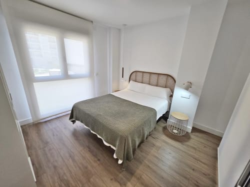 Dormitorio luminoso en apartamento BR Silgar Sanxenxo con cama de ratán, mesita blanca y puertas al balcón