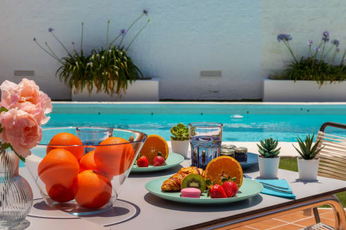 Poolside breakfast setup at Ayma Casa Nísperos with fresh oranges, pastries, and pink flowers overlooking the turquoise pool.