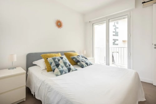 Chambre moderne avec balcon vue mer, lit capitonné gris, coussins géométriques, linge blanc, appartement côtier