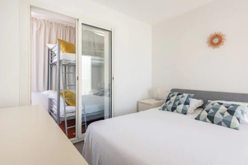 Chambre moderne avec lit queen-size, lits superposés visibles par la porte vitrée, coussins géométriques et horloge murale en cuivre chez You Camp Immobilier