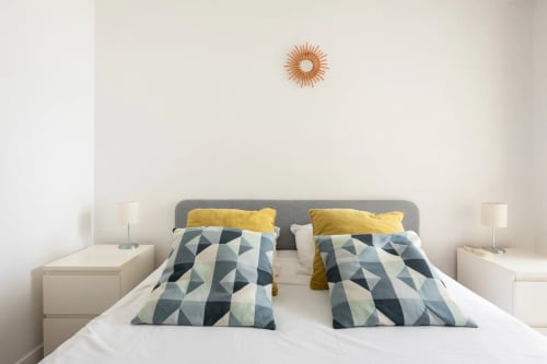 Chambre moderne avec tête de lit grise, coussins à motifs géométriques, accents jaunes, literie blanche et miroir soleil en cuivre