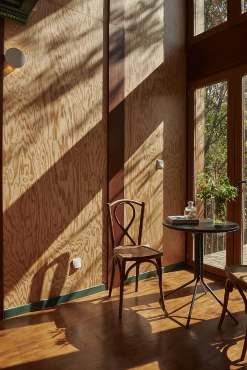 Chambre lumineuse aux Micro Palaces B&B à Vaulx avec chaise en bois courbé, petite table et murs en contreplaqué projetant des ombres géométriques.