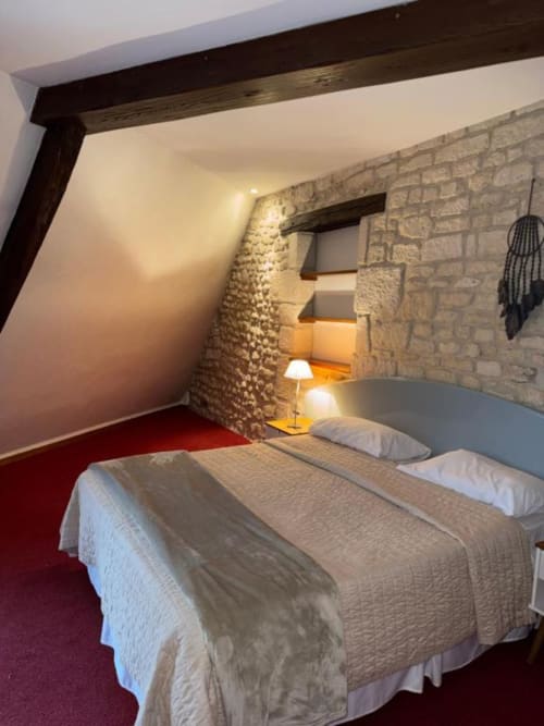 Chambre mansardée au Château des Saveurs avec deux lits king-size, poutres apparentes, murs en pierre, tapis rouge et éclairage chaleureux
