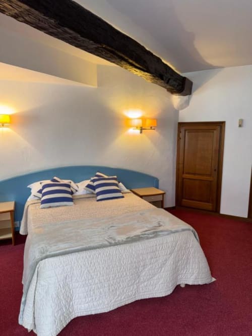 Chambre double douillette au Château des Saveurs avec tête de lit bleue, oreillers rayés, poutre en bois apparent et moquette rouge