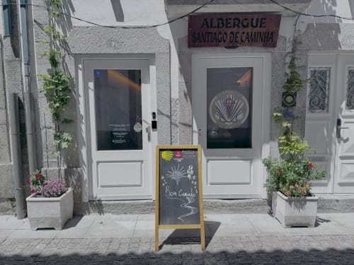 Albergue Santiago de Caminha hostel entrance in Caminha with white doors, scallop shell signage, and flowering planters