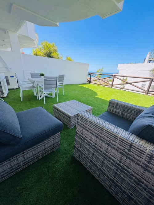 Amplia terraza con vistas al mar, césped artificial, zona de descanso, mesa de comedor y sombrillas blancas con vista a la
