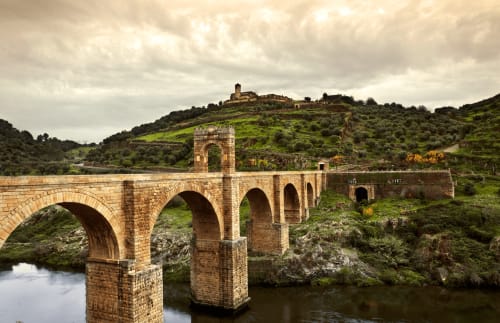 Puente medieval de piedra atravesando río con pueblo en loma al fondo, Cáceres España