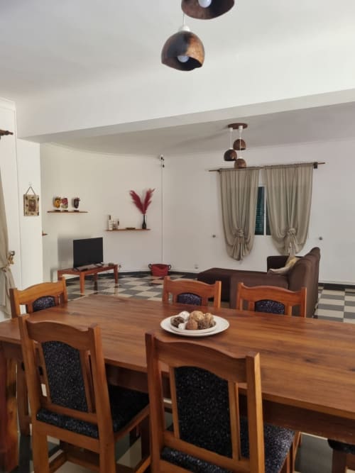 Área de jantar da pousada Casa Amarela em Equador Houses, São Tomé, com mesa de madeira, luminárias pendentes marrom e vista para a sala de estar