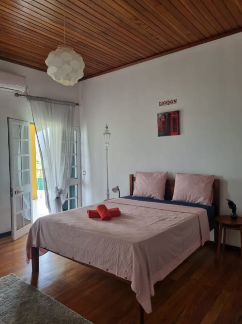 Suíte espaçosa da Casa Amarela em Equador Houses, São Tomé, com teto de madeira, roupa de cama rosa e vista para o jardim