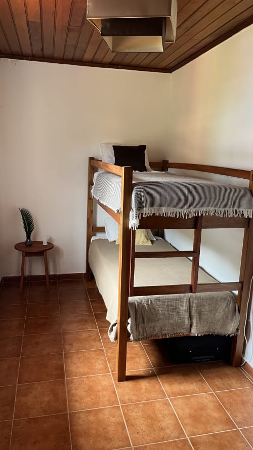 Beliche de madeira com roupa de cama listrada no dormitório Equador Houses, São Tomé, com piso de azulejo terracota e teto de madeira.