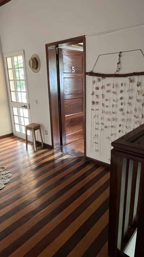 Quarto 5 nas Casas Equador em São Tomé, com piso de madeira listrada, porta de madeira escura, janela e painel decorativo na parede