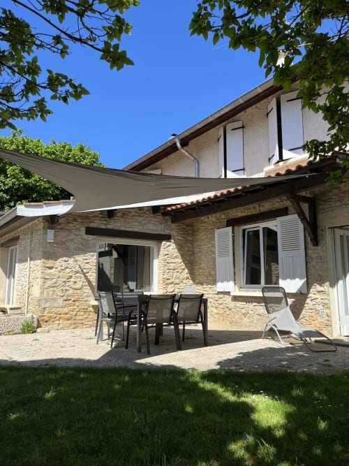 Terrasse du gîte Le Saint-Hilaire avec façade en pierre, voile d'ombrage, table de repas et vues sur les montagnes à Saint-Hilaire-de-Brens