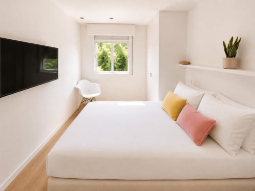 Dormitorio moderno en apartamento de Baiona con cama blanca, almohadas amarillas y rosas, televisor de pantalla plana, ventana con vistas a la ciudad y maceta