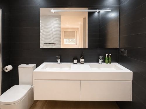 Baño moderno con lavabo doble, paredes de azulejo negro y espejo iluminado en el apartamento Holibai de Baiona