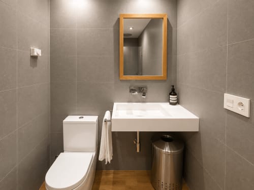 Baño moderno en apartamento Campaiña, Baiona con lavamanos blanco, inodoro, espejo con marco de madera y paredes con azulejos grises