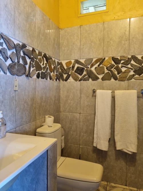 Banheiro da suíte Vila Paraíso em Equador Houses, São Tomé, com azulejos bege, friso decorativo em pedra, vaso sanitário, pia e toalha