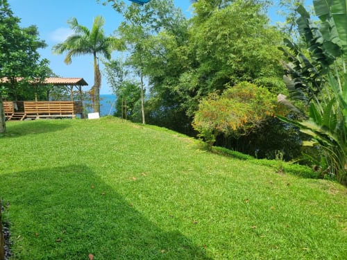 Jardim exuberante com vista para o mar na Ecuador Houses, São Tomé. Palmeiras e um pavilhão de madeira com vista para o oceano.